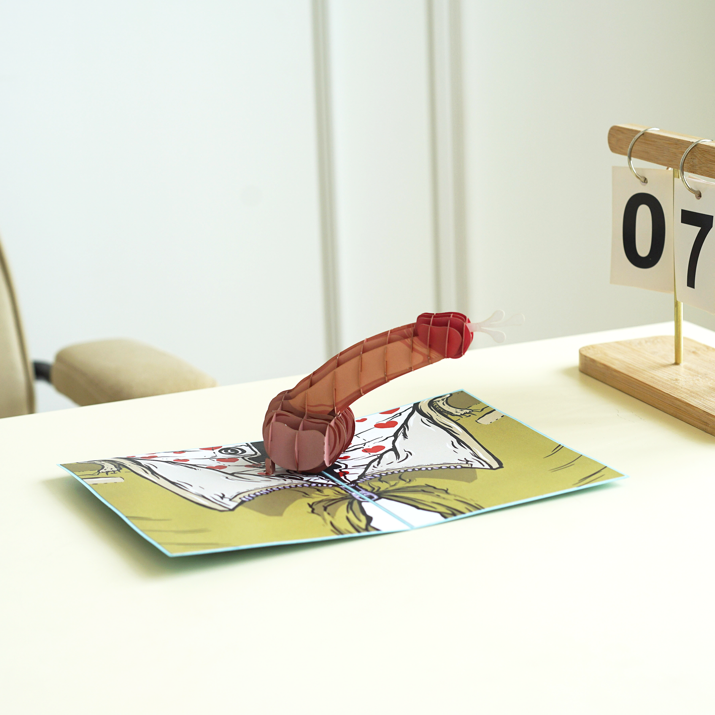 Pop-up book with a red creature on a white surface, calendar in the background