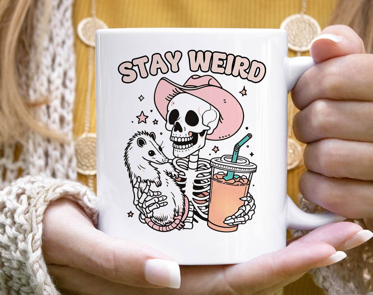 Mug with skeleton and opossum design and 'Stay Weird' text held by a person.