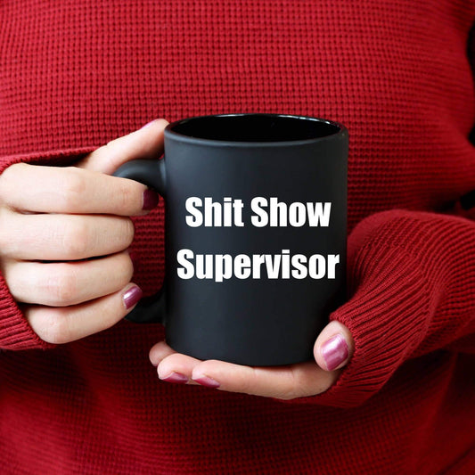 Person holding a black mug with 'Shit Show Supervisor' text, wearing a red sweater.