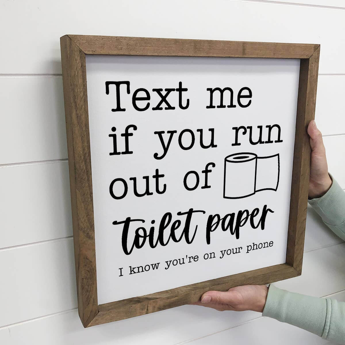 Wooden-framed sign with humorous text about toilet paper, held against a white wall.
