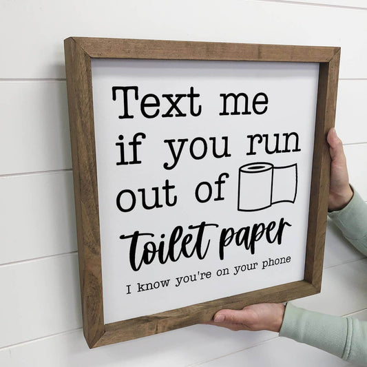 Wooden-framed sign with humorous text about toilet paper, held against a white wall.