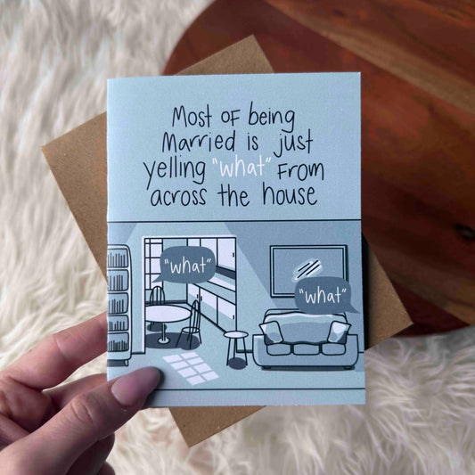 Card with humorous illustration and text about married life held by a hand on a soft surface.