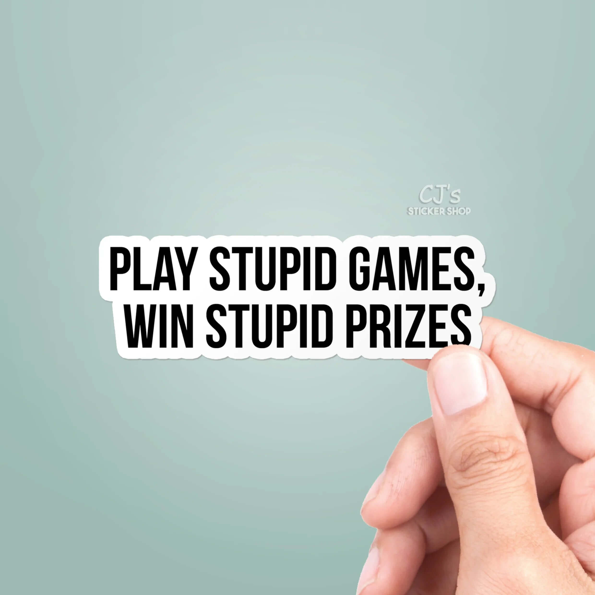 Hand holding a sticker with text 'Play Stupid Games, Win Stupid Prizes' on a light gray background.