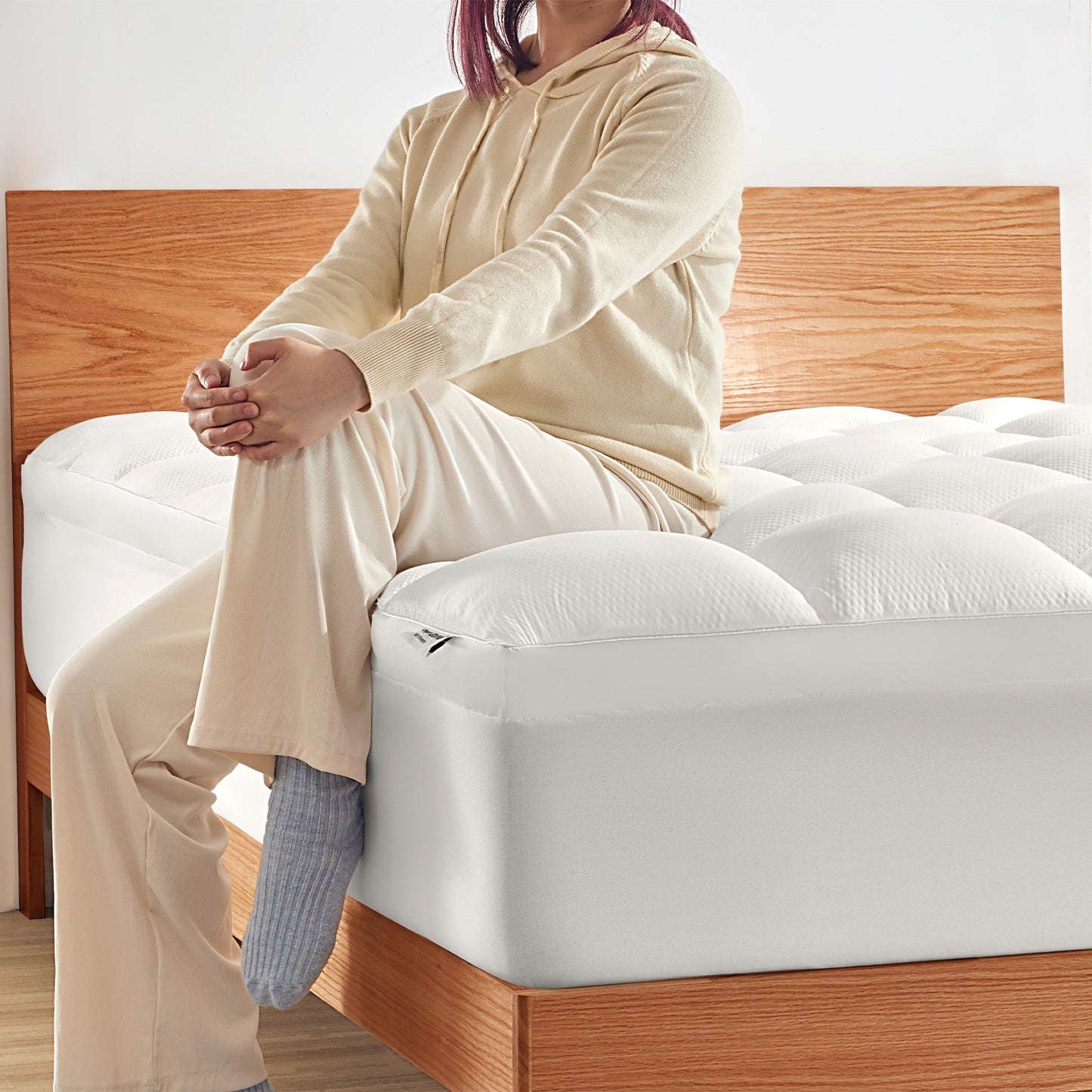 Person sitting on a bed with a mattress topper, wearing a beige outfit.