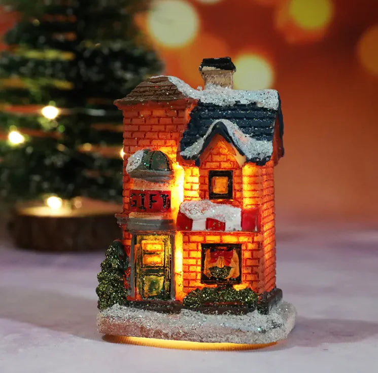 Decorative Christmas house with glowing interior, snow on roof, and 'Gift' sign, against a blurred Christmas tree background.