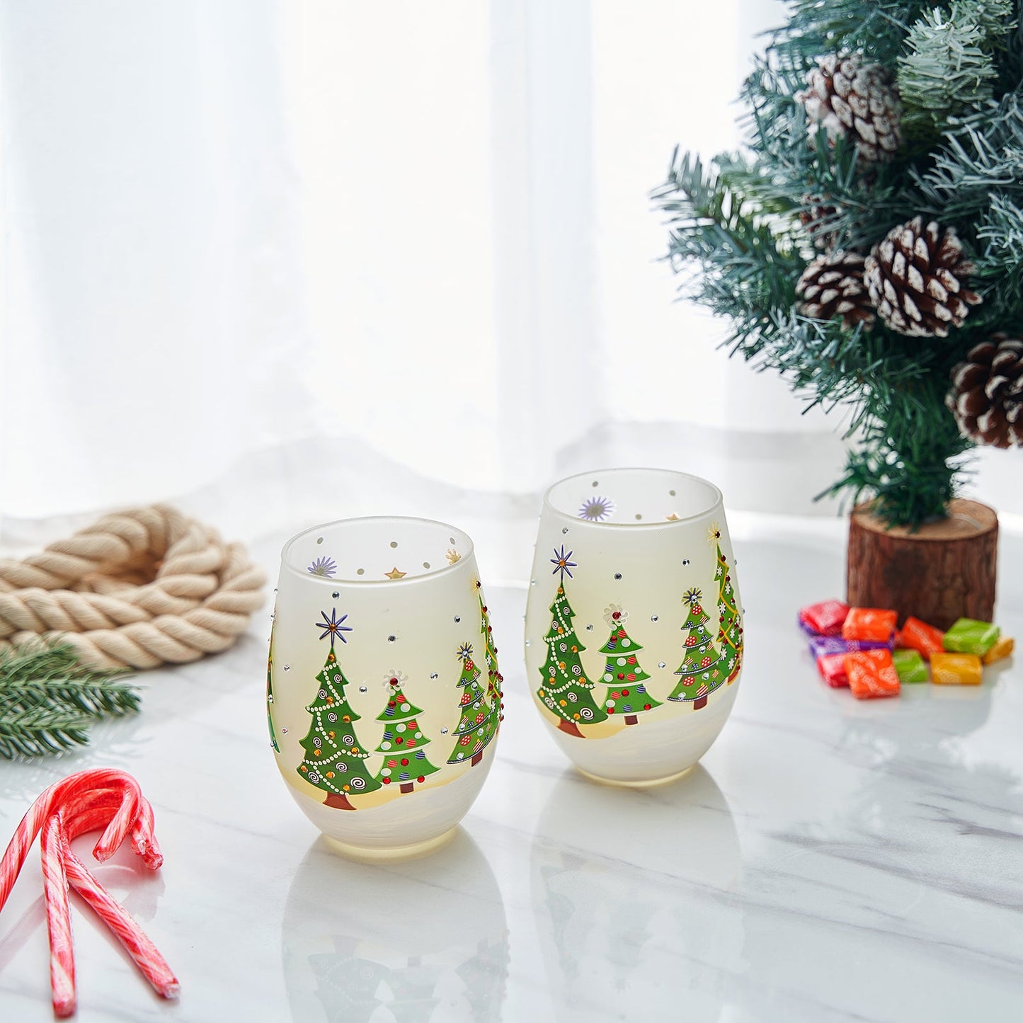 Two Christmas-themed stemless wine glasses on a table with a decorated tree in the background.