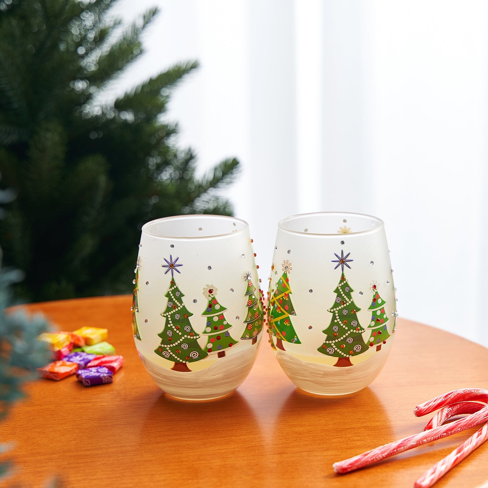 Two stemless wine glasses with Christmas tree designs on a wooden table with candy canes and wrapped gifts.