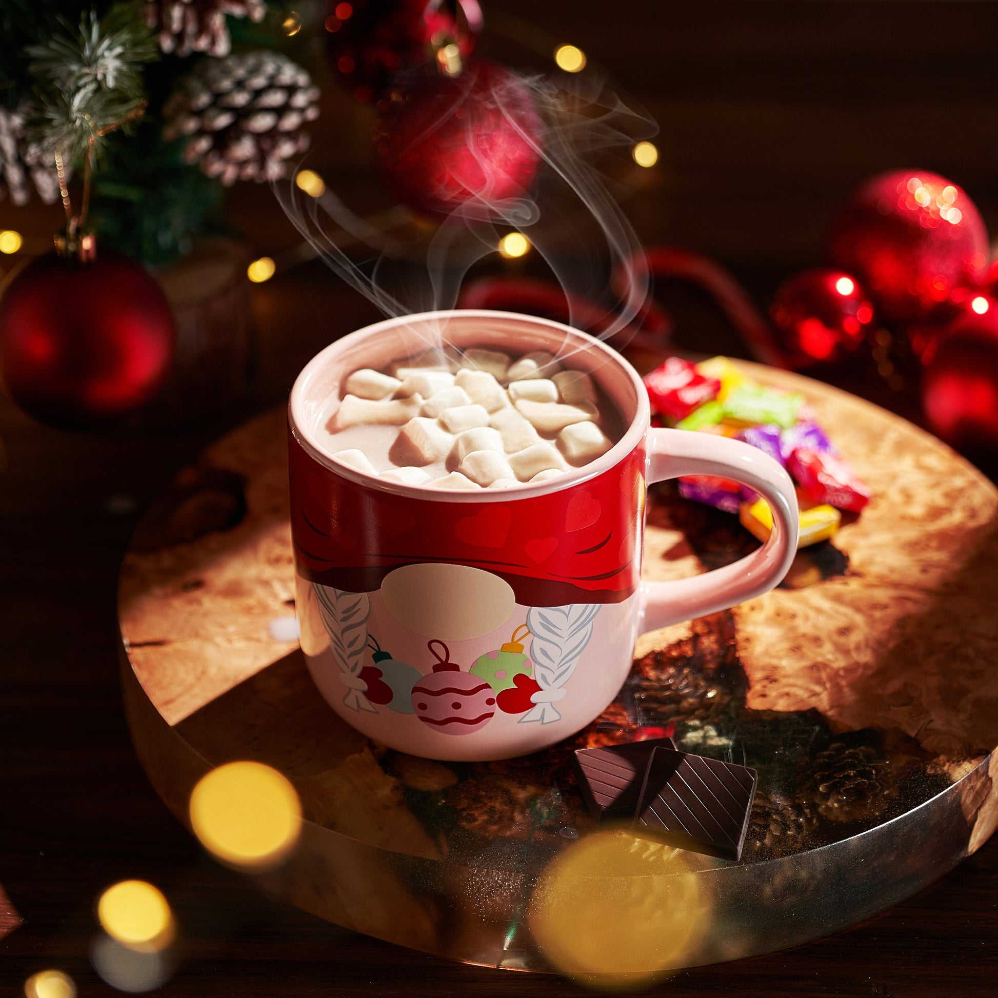 Red mug with cartoon character filled with hot chocolate and marshmallows, surrounded by Christmas decorations.