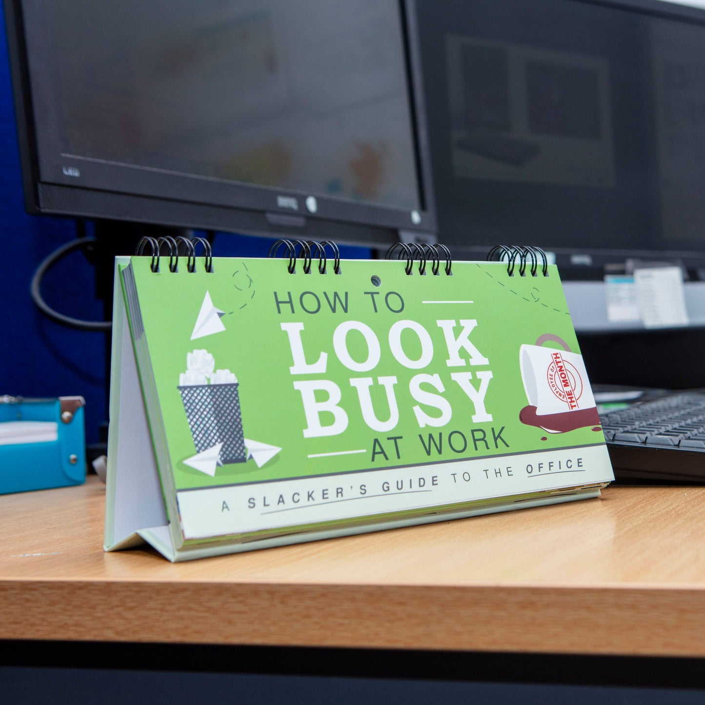 Desk calendar titled 'How to Look Busy at Work' on a desk with computer monitors in the background