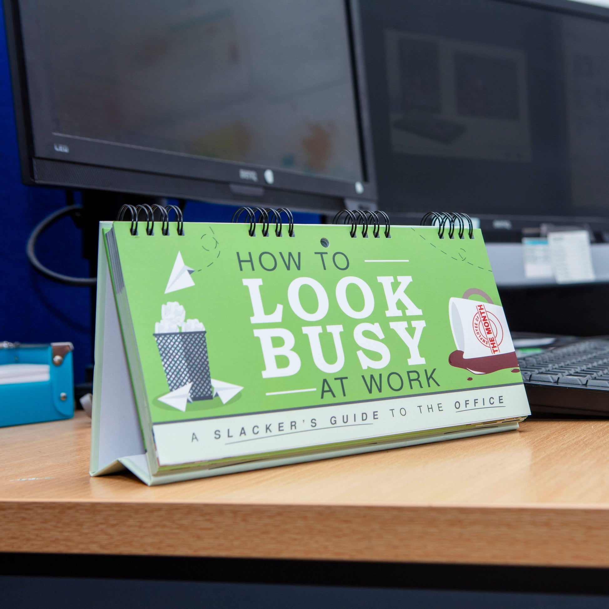 Desk calendar titled 'How to Look Busy at Work' on a desk with computer monitors in the background