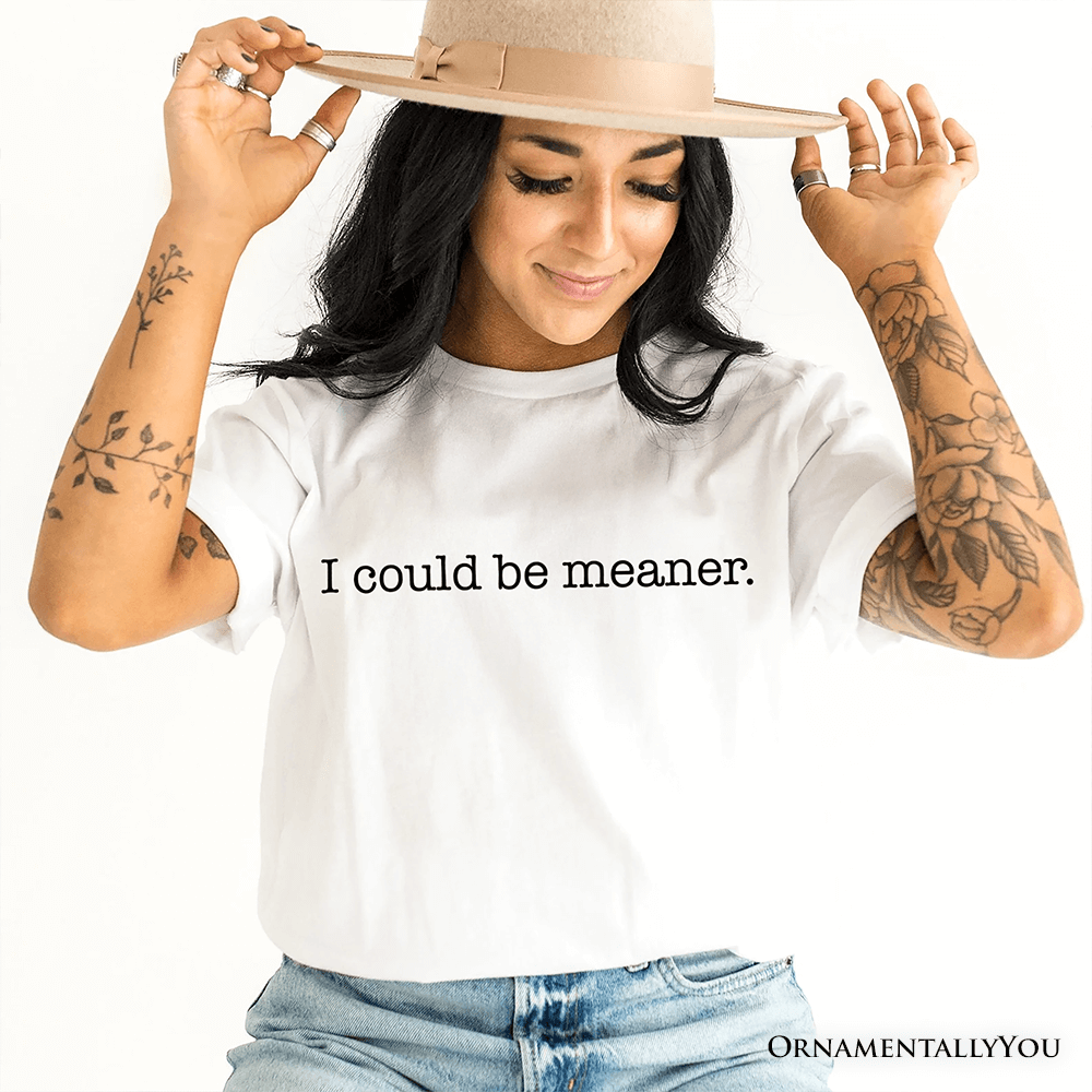 Woman wearing a white t-shirt with 'I could be meaner.' text, holding a beige hat.