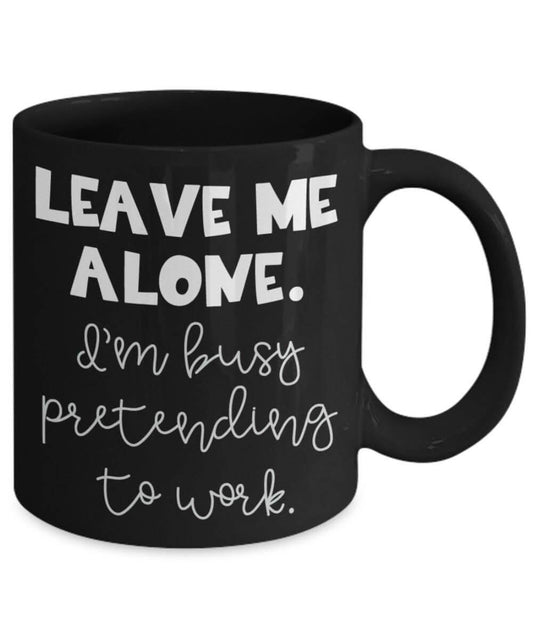 Black mug with white text 'LEAVE ME ALONE. I'm busy pretending to work.' on a white background