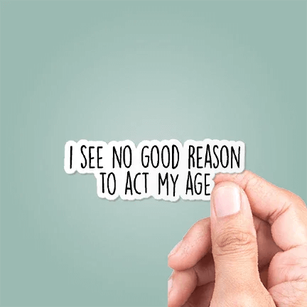 Hand holding a sticker with text 'I see no good reason to act my age' against a light blue background