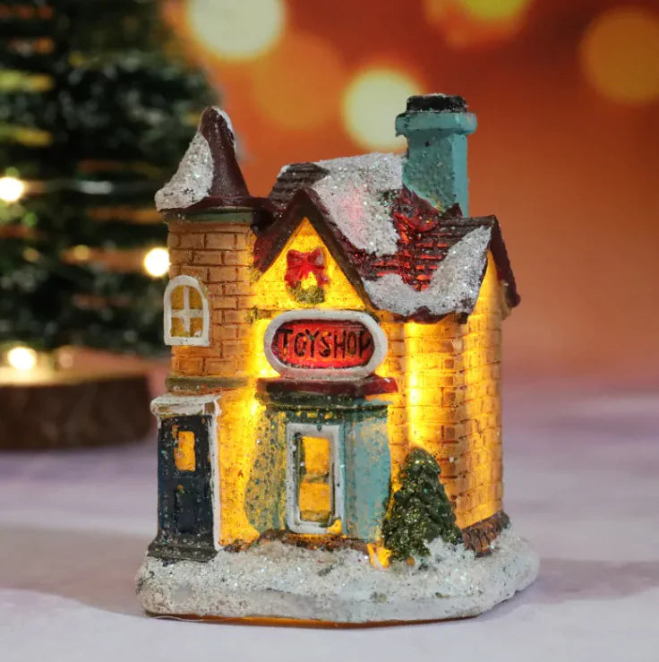 Decorative toy shop house with glowing interior, snow on roof, and 'Toy Shop' sign, blurred Christmas tree in background.