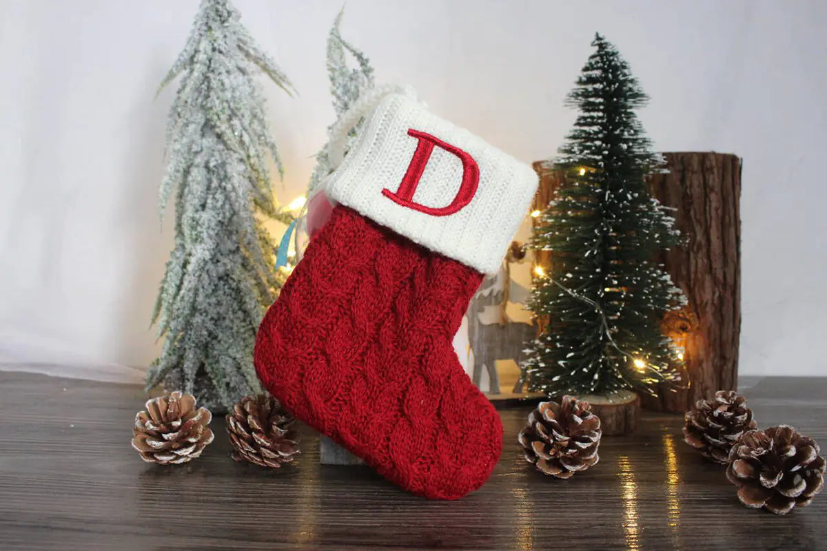 Red Christmas stocking with white cuff featuring a red letter 'D', surrounded by decorative trees and pinecones on a wooden surface.